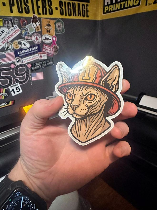 Hand holding a full color die cut sticker of a cat wearing a firefighter helmet, in the Hynds Printing shop